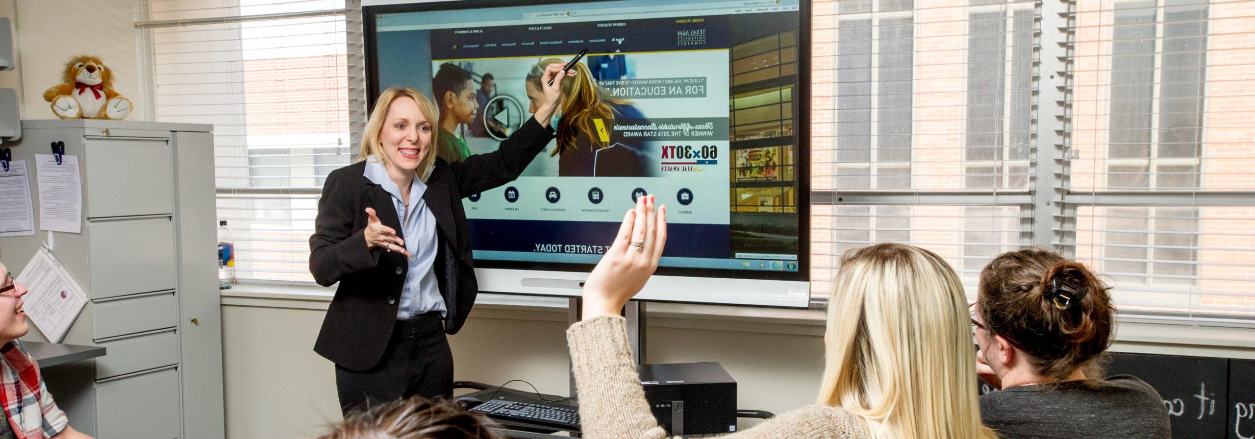 woman in professional attire teaching young adults in classroom.